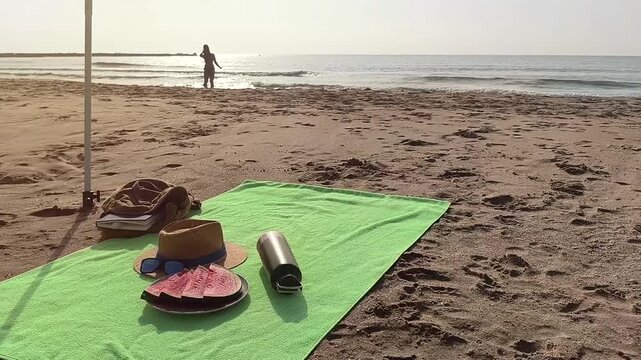 Beach towel spread on golden sand with a plate of freshly sliced watermelon, a sun hat with sunglasses, and a reusable water bottle. In the background, the silhouette of a woman enjoys the sea