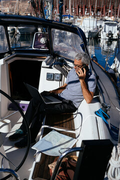Senior captain making phone call while sitting on sailboat with laptop