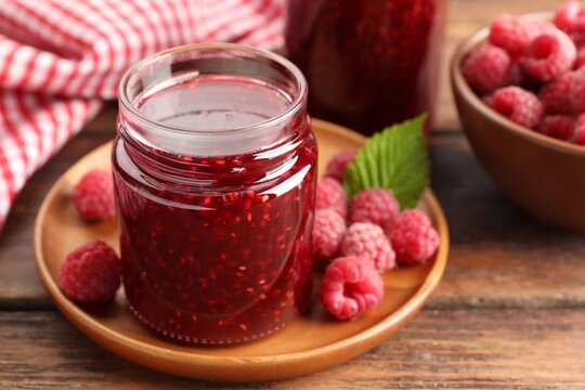 Tasty raspberry jam in jars and fresh berries on wooden table, closeup