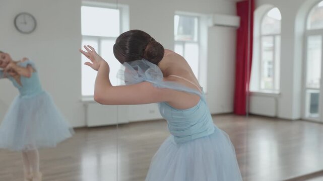 Young elegant woman in a light blue tutu dress practicing classical ballet movements, gracefully performing arabesques and pirouettes in a spacious, sunlit dance studio with mirrors