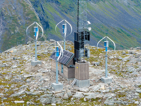 Aerial view of vertical axis wind turbines, a wooden cabin with solar panels, and a communication tower on the rocky terrain of Trollstigen in Andalsnes, More og Romsdal, Norway.
