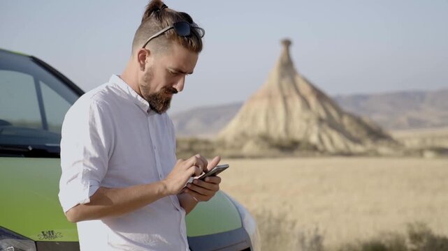 Caucasian man using cellphone outdoors in a desert standing next to a green van, sending online text messages. Copy space footage.