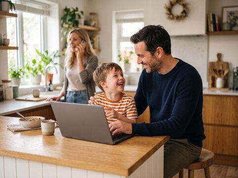 Caring Father Helping His Smiling Young Son Use a Laptop at the Kitchen Counter While Mother Talks on the Phone in a Warm Modern Family Lifestyle Scene