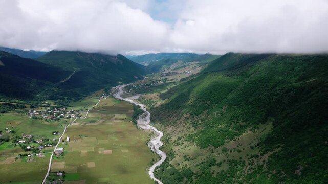 Cinematic drone flight above Zhabeshi village in Svaneti, Georgia. Stunning aerial view of green mountain valley, alpine river, dramatic Caucasus peaks and clouds, for travel, documentary, meditation 