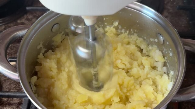 Electric mixer mashes yellow potatoes in a steel pot on a home stove. Homemade cooking, dinner preparation, comfort food