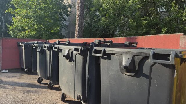Row of black plastic trash bins with wheels near a red wall outdoors. Urban waste collection, garbage containers, street cleanup