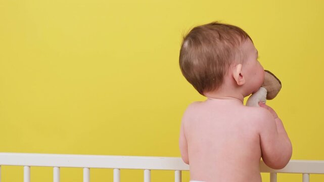 Baby chewing on a plush toy while standing in a white crib. Cute toddler playing with a stuffed animal on a yellow background. Childhood development, nursery concept and care activity.