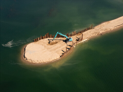 Aerial view of an excavator working on a sandy peninsula at Lampedziai Beach, surrounded by green water and metal sheet piling in Kaunas, Kauno apskritis, Lithuania.