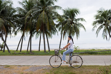 Fototapeta premium Asian Woman Cycling on Coastal Road Lined with Coconut Trees,A candid, full-body photograph of a young Asian woman in her late 20s riding a vintage cruiser bicycle with a front basket along a quiet.