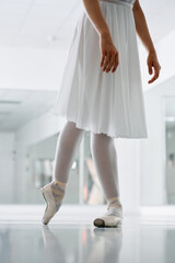Cropped low-angle view of a female ballet dancer in a white dress and point shoes practicing elegant footwork in a bright modern dance studio. © Oksana