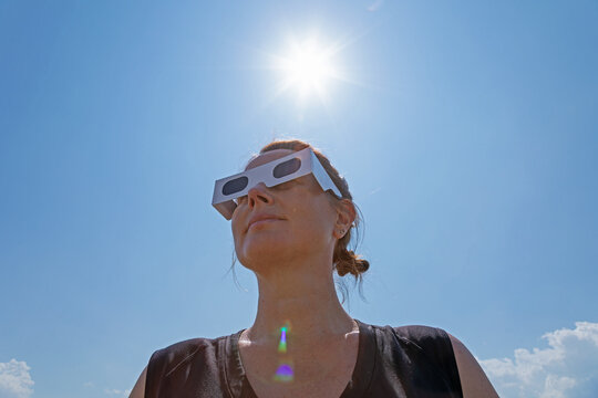 woman wearing protective solar eclipse glasses with the bright sun positioned directly above her in a clear blue sky, preparing to observe the upcoming solar eclipse on August 12, 2026.