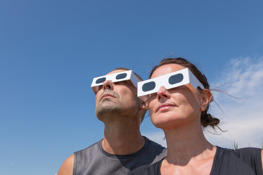 Low-angle close-up view of a man and a woman wearing protective paper glasses, looking up into a clear blue sky to observe the upcoming solar eclipse on August 12, 2026.