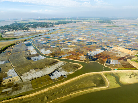 Aerial view of salt pans in the coastal region of Cox's Bazar, featuring geometric evaporation ponds, winding waterways, and distant wind turbines. Cox's Bazar, Chittagong Division, Bangladesh.