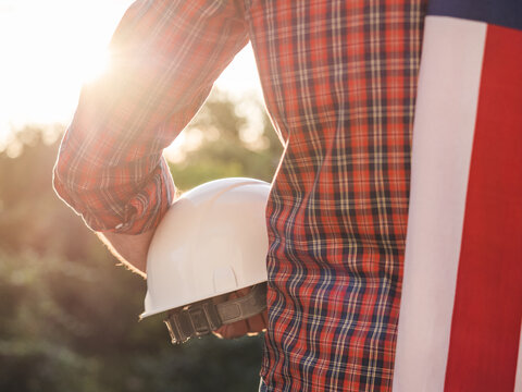 Construction worker holds a white hard hat with an American flag draped over his shoulder. Symbolizes American workers or Labor Day. Concept of pride in construction profession and American dream