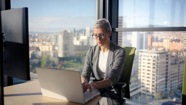Middle aged businesswoman types on a laptop beside a large window with a panoramic city skyline, focused on daily tasks in a modern corporate office during daylight.