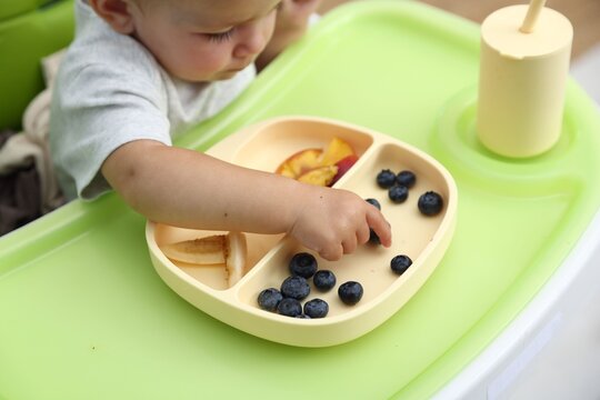 Baby tableware. Cute little child eating food in high chair indoors, closeup
