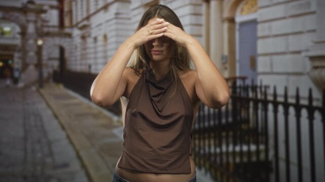 Young woman shields her eyes with hands on a city street by iron railings and stone building, wearing a sleeveless top; privacy vulnerability.