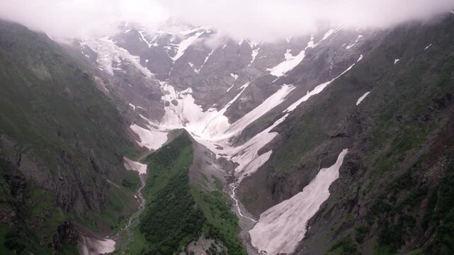 Aerial drone flight over a glacier valley in Svaneti, Georgia. Mountain river, lush green slopes, snow-covered peaks, glaciers and clouds in breathtaking summer Caucasus landscape