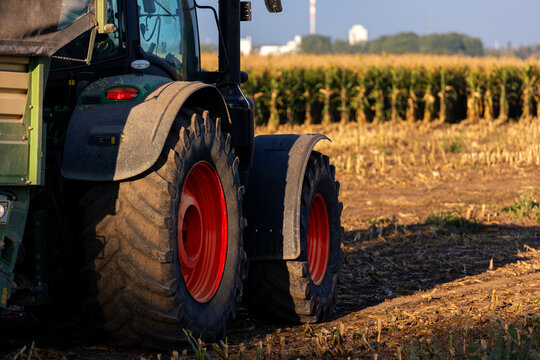 Close view of tractor wheels beside harvested cornfield in warm evening sunlight. Heavy agricultural machinery reflects maize harvesting, rural work, mechanization, farming, and seasonal production