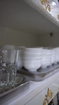 Close up of clean glass tumblers and stacked white ceramic bowls on metal trays inside a classic kitchen cabinet, slow motion vertical video.