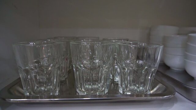 Slow motion camera panning of clean empty glass tumblers arranged on a stainless steel tray inside a kitchen cabinet, horizontal video.