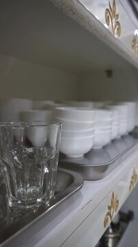 Close up of clean glass tumblers and stacked white ceramic bowls on metal trays inside a classic kitchen cabinet, slow motion vertical video.