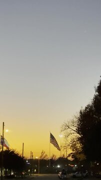 American flag, orange sunset, patriotic scene, flag at dusk, evening sky, national symbol, golden horizon, waving flag, silhouetted trees, street lights, twilight landscape, outdoor ceremony, peaceful