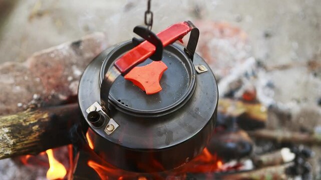Black metal kettle with red lid handle boiling over outdoor bonfire in natural rustic setting