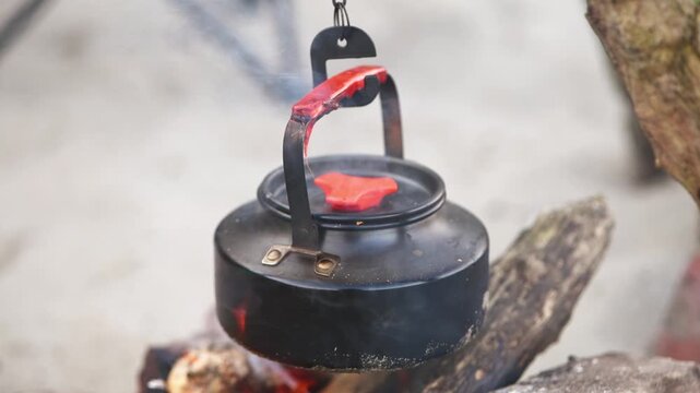 Black kettle heating over outdoor bonfire hanging from metal hook in natural setting