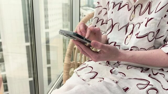 Person using smartphone near a window in a bright apartment. Handheld tapping on a mobile device with city view background. Remote communication and online work concept..