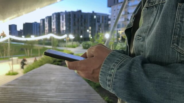 Phone held steady, a man drafts a text message beneath a canopy in a modern residential complex at dusk. Buildings glow softly behind him as evening settles over the city park. He pauses