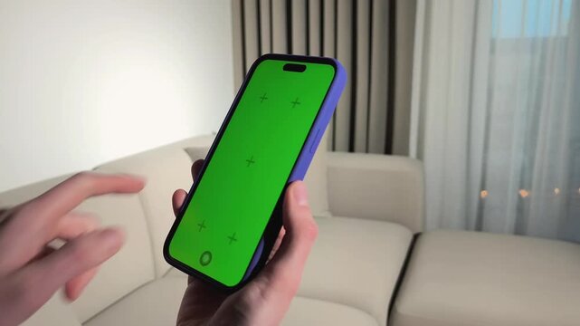 Hand holds smartphone with green screen in close up. Finger moves toward the vertical display on a sofa. Mobile mockup for app design and advertising..