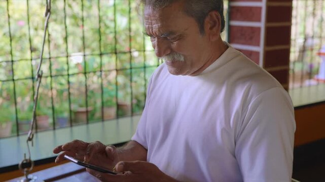 Daylight indoor close shot of healthy, elderly Indian Asian man with grey hair sitting on wooden swing on the veranda of traditional house, using mobile phone to type text message or use social media