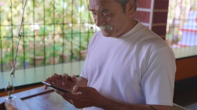 Daylight indoor close shot of healthy, elderly Indian Asian man with grey hair sitting on wooden swing on the veranda of traditional house, using mobile phone to type text message or use social media