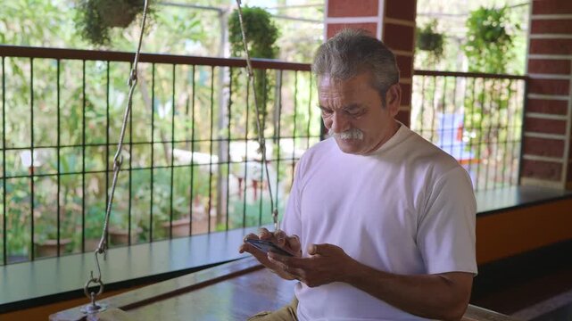 Daylight indoor mid shot of healthy, elderly Indian Asian man with grey hair sitting on a wooden swing on veranda of traditional house, using mobile phone to type a text message or to use social media