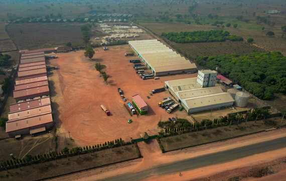 Aerial view of an industrial warehouse complex with storage buildings, parked trucks, and a dirt yard near the Abuja - Kaduna - Zaria Express Way, Kaduna, Kaduna, Nigeria.