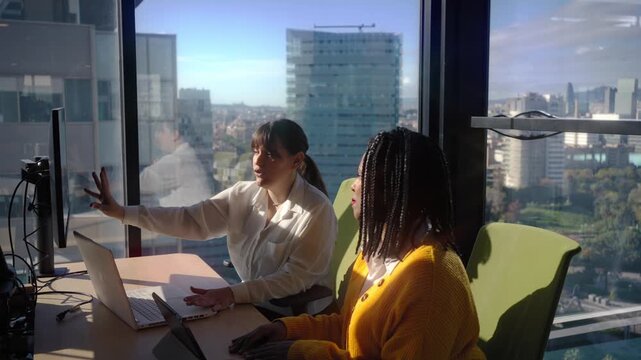 Two female colleagues review a project at a desk, pointing at a monitor and laptop in a bright office with city views, conveying teamwork, guidance, and productive collaboration during a meeting.