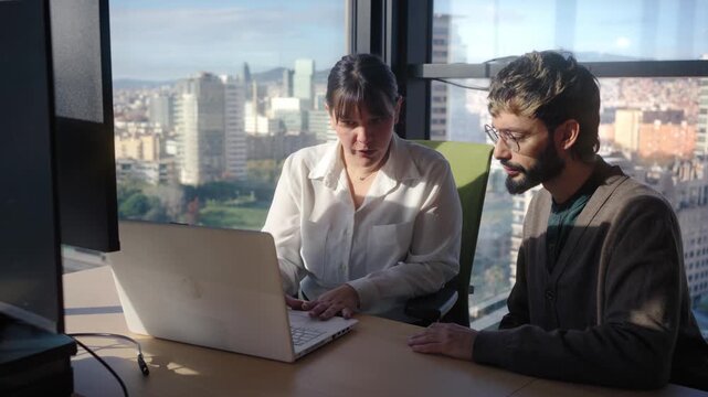 Male and female colleagues having a focused conversation beside a laptop in a sunlit high-rise office. Corporate scene conveying consultation, problem solving and professional collaboration.