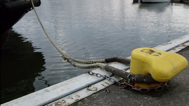 Close-up of mooring ropes secured to a yellow harbor bollard on a pier with calm water in the background. Static detail of maritime infrastructure used for docking boats and ships in a modern marina