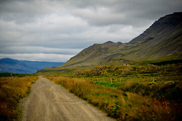 Naklejka premium Unpaved gravel road stretching through Icelandic countryside landscape with mountains under cloudy sky