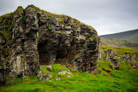 Basalt Rock Formation with Cave Opening and Green Hills Under Cloudy Sky in Iceland