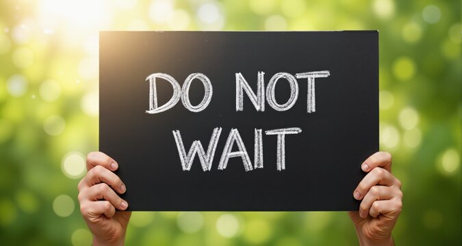 Hands hold blackboard sign with chalk text do not wait. Message encourages immediate action. Soft natural green bokeh background with bright sunlight.
