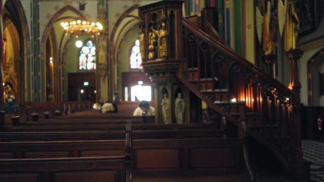 Blurred figures sit in ornate catholic church with wooden pews and intricate staircase under soft lighting, creating a peaceful, reflective atmosphere indoors.