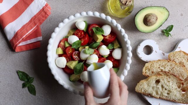 Cooking avocado caprese salad. Olive oil added to a salad with cherry tomatoes, mozzarella cheese, basil in a white bowl on a beige background. Mediterranean healthy food concept. Horizontal video.