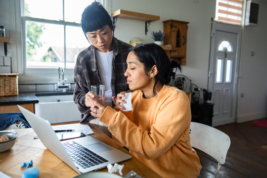 Two adults caring for a sick partner at a home kitchen table with laptop