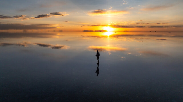 Aerial view of a person standing on the Uyuni Salt Flat at sunset with a mirror reflection on the water surface in Potosi Department, Bolivia.