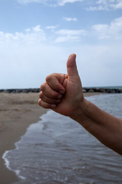 Primo piano di una mano umana che fa il gesto del pollice in su in segno di approvazione sullo sfondo di una spiaggia con mare calmo