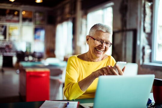 Smiling senior woman using smartphone and laptop at cafe