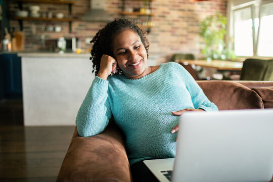 Smiling pregnant woman using laptop on sofa at home
