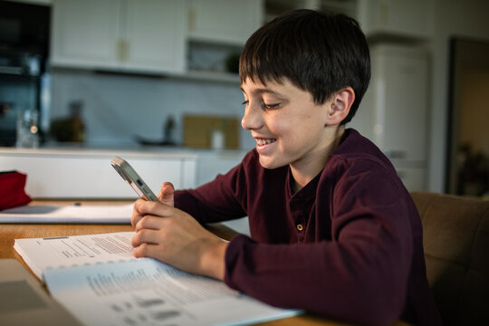 Teen boy using smartphone while doing homework at kitchen table